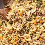 Egg noodles mixed with ground beef, peas, carrots, and a creamy sauce, garnished with chopped parsley, in a pan with a wooden spoon.