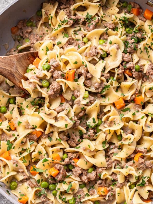 Egg noodles mixed with ground beef, peas, carrots, and a creamy sauce, garnished with chopped parsley, in a pan with a wooden spoon.