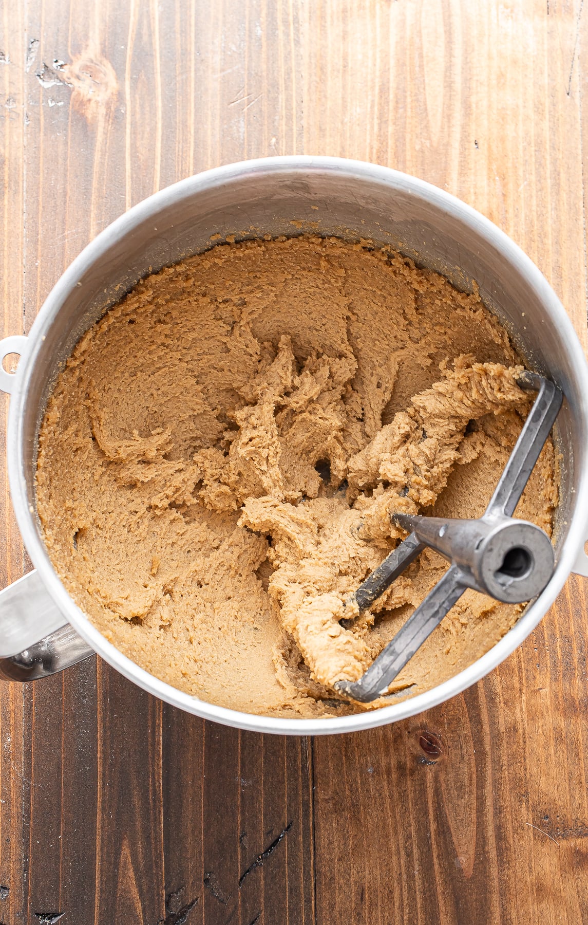 A metal mixing bowl with light brown cookie dough and a paddle attachment sits on a wooden surface.