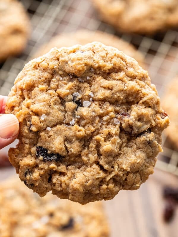 A hand holds a large, round oatmeal cookie with visible oats and raisins. Other cookies are blurred in the background on a cooling rack.