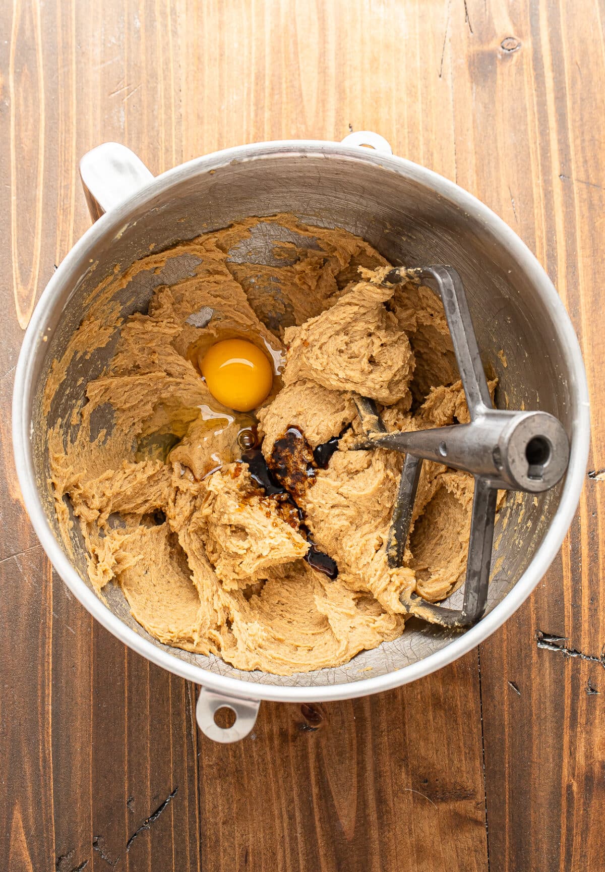 A mixing bowl with cookie dough, a raw egg, and vanilla extract, partially mixed with a stand mixer paddle attachment on a wooden surface.