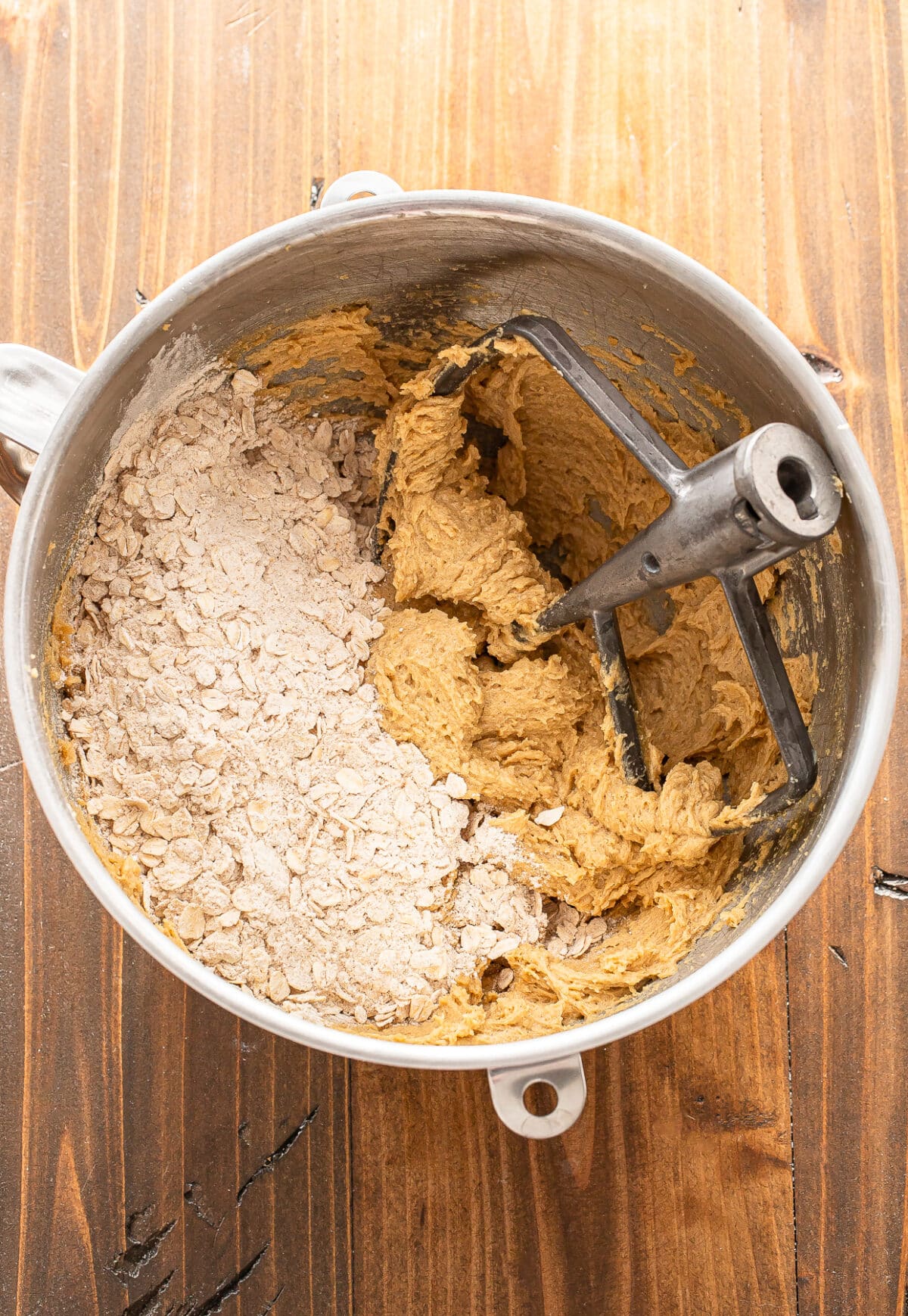 A mixing bowl containing partially mixed cookie dough, with oats and flour on one side and creamy dough on the other. A paddle attachment is in the bowl, and the bowl sits on a wooden surface.
