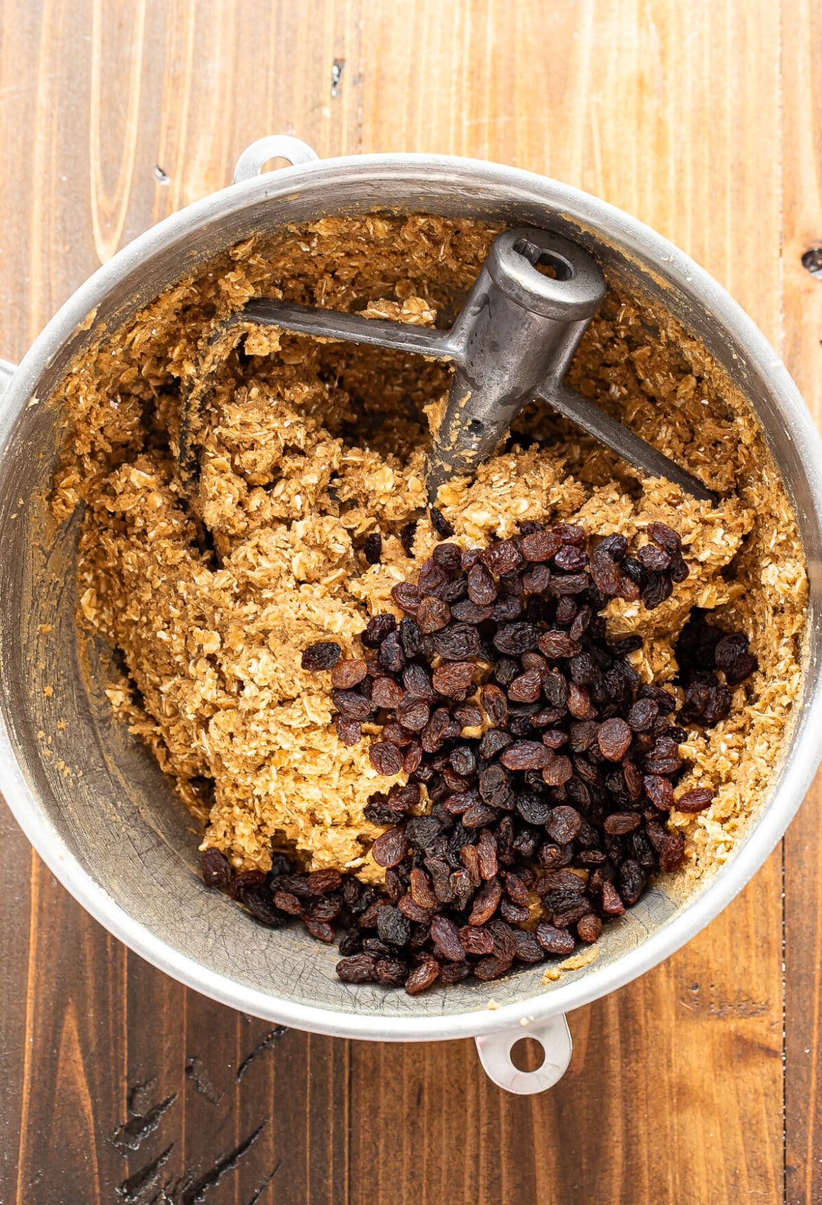 A mixing bowl with oatmeal cookie dough, a paddle attachment, and a pile of raisins on top, all set on a wooden surface.