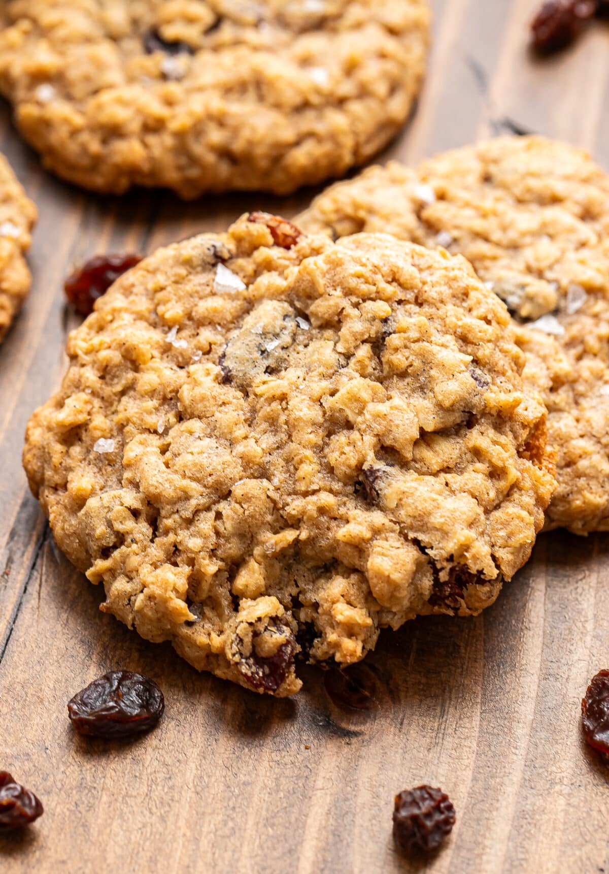 Three oatmeal raisin cookies on a wooden surface with several scattered raisins nearby. The cookies have a chewy texture with visible oats and raisins.