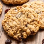 Three oatmeal raisin cookies on a wooden surface with several scattered raisins nearby. The cookies have a chewy texture with visible oats and raisins.