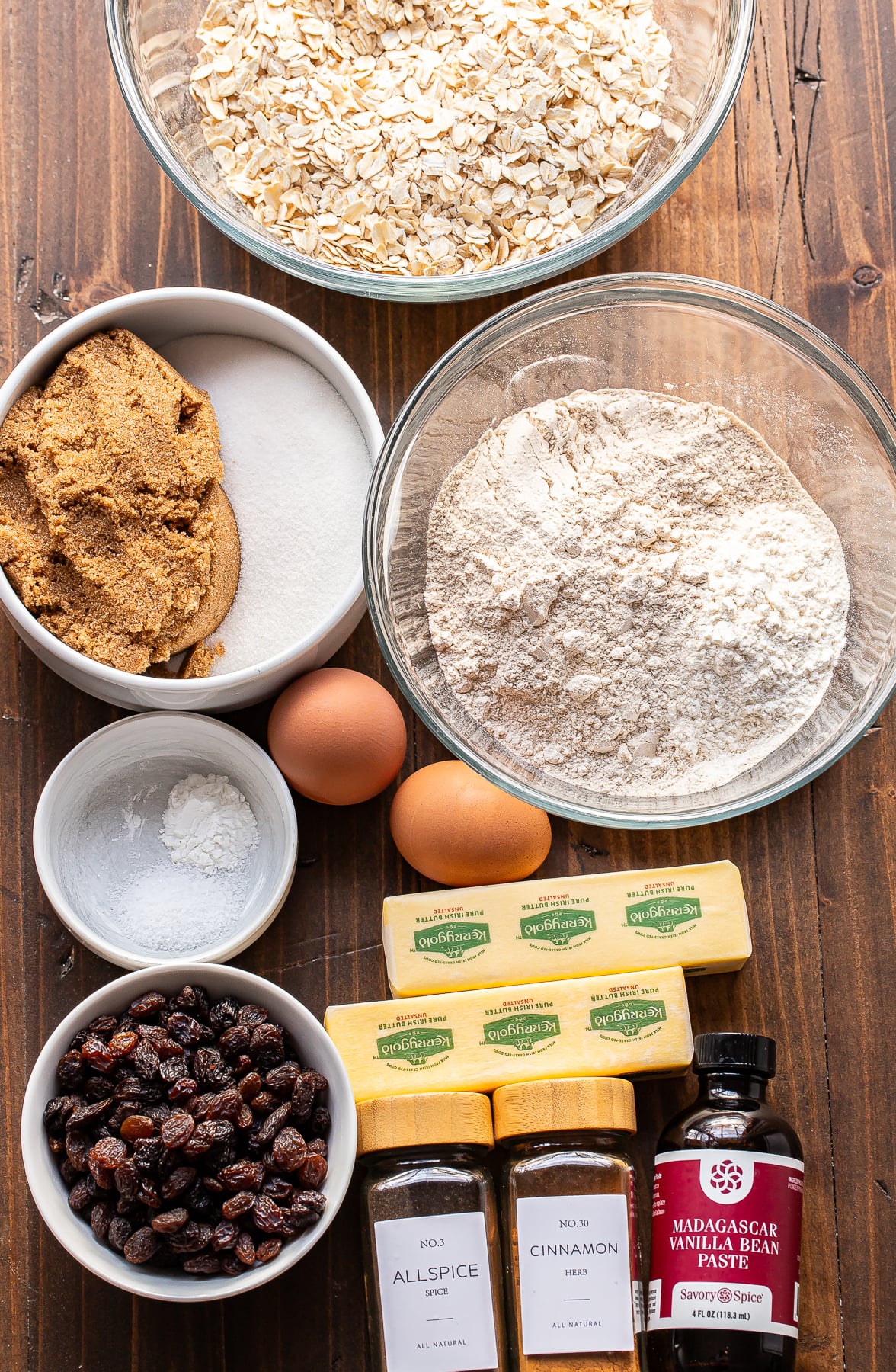 Bowls with oats, flour, brown and white sugar, baking powder, two eggs, butter sticks, raisins, jars of allspice and cinnamon, and a bottle of vanilla extract arranged on a wooden table.
