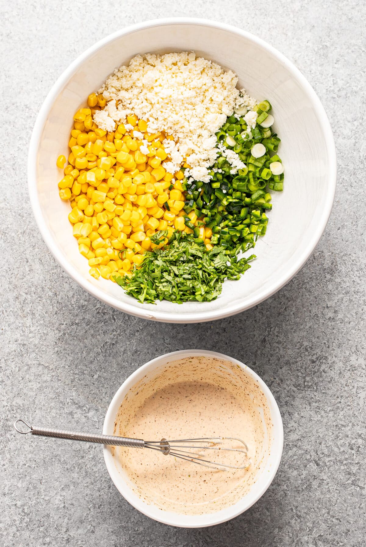A large bowl with corn, feta cheese, chopped green onions, jalapeños, and cilantro. Below it, a small bowl of creamy dressing with a whisk. Both are on a light gray surface.