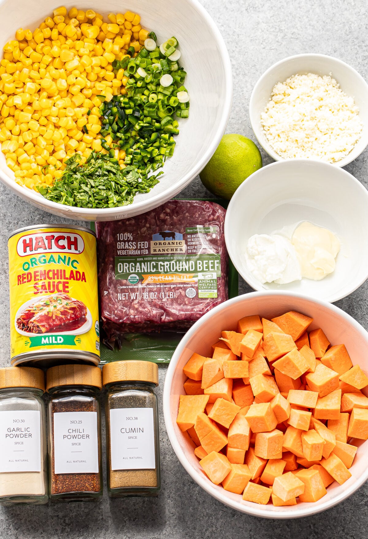 Bowls of chopped sweet potatoes, corn, green onions, crumbled cheese, and sauces, with packages of ground beef, enchilada sauce, and bottles of garlic powder, chili powder, and cumin on a gray surface.