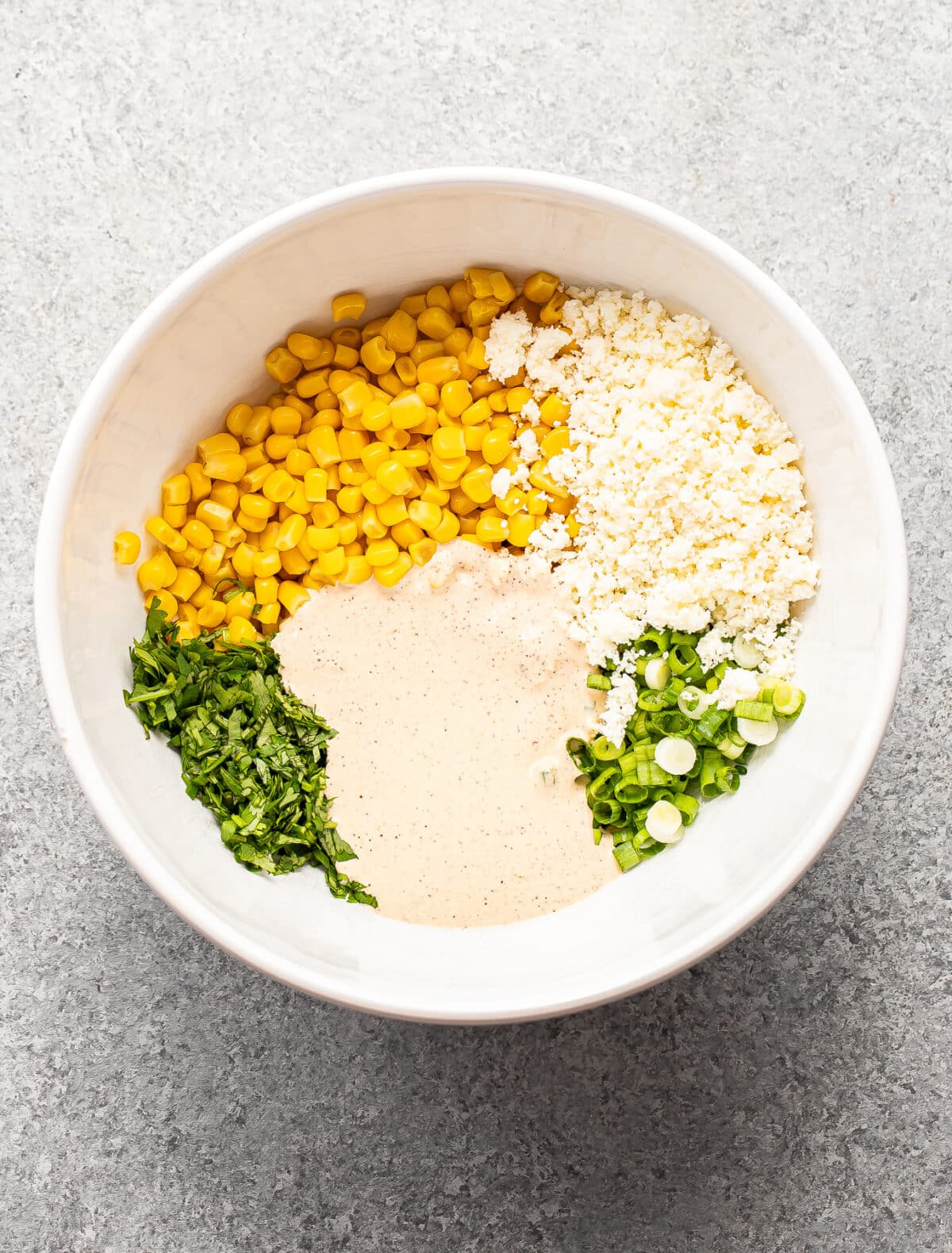 A white bowl with corn, crumbled cheese, sliced green onions, chopped cilantro, and creamy dressing, all separated in sections on a gray surface.