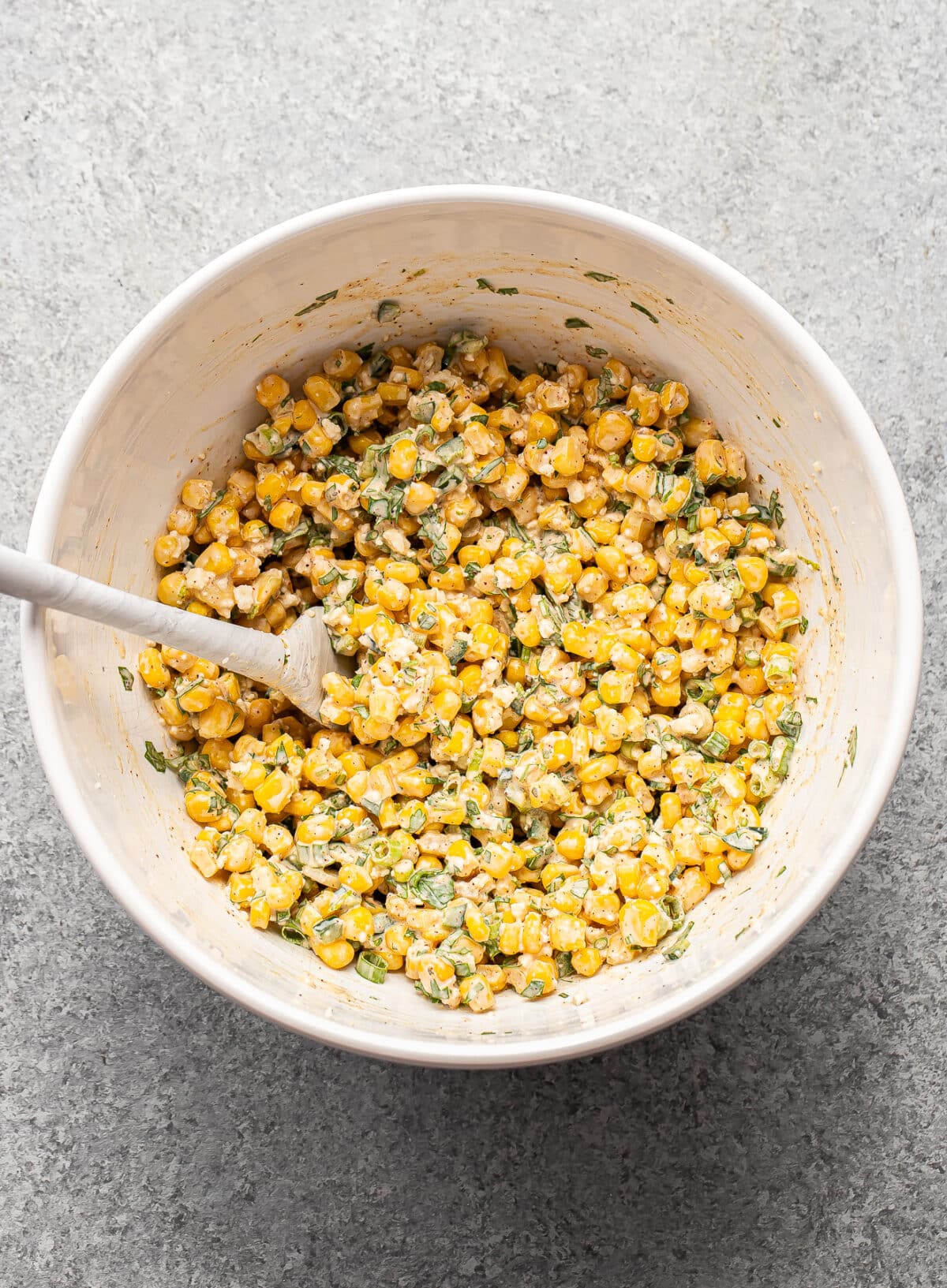 A white bowl filled with a creamy corn salad mixed with chopped green herbs and a white spoon resting inside, set on a gray textured surface.