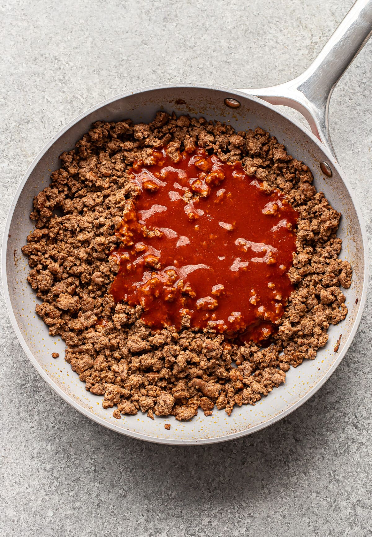 Cooked ground beef in a skillet with red tomato sauce poured in the center, ready to be mixed together. The pan is placed on a light gray countertop.