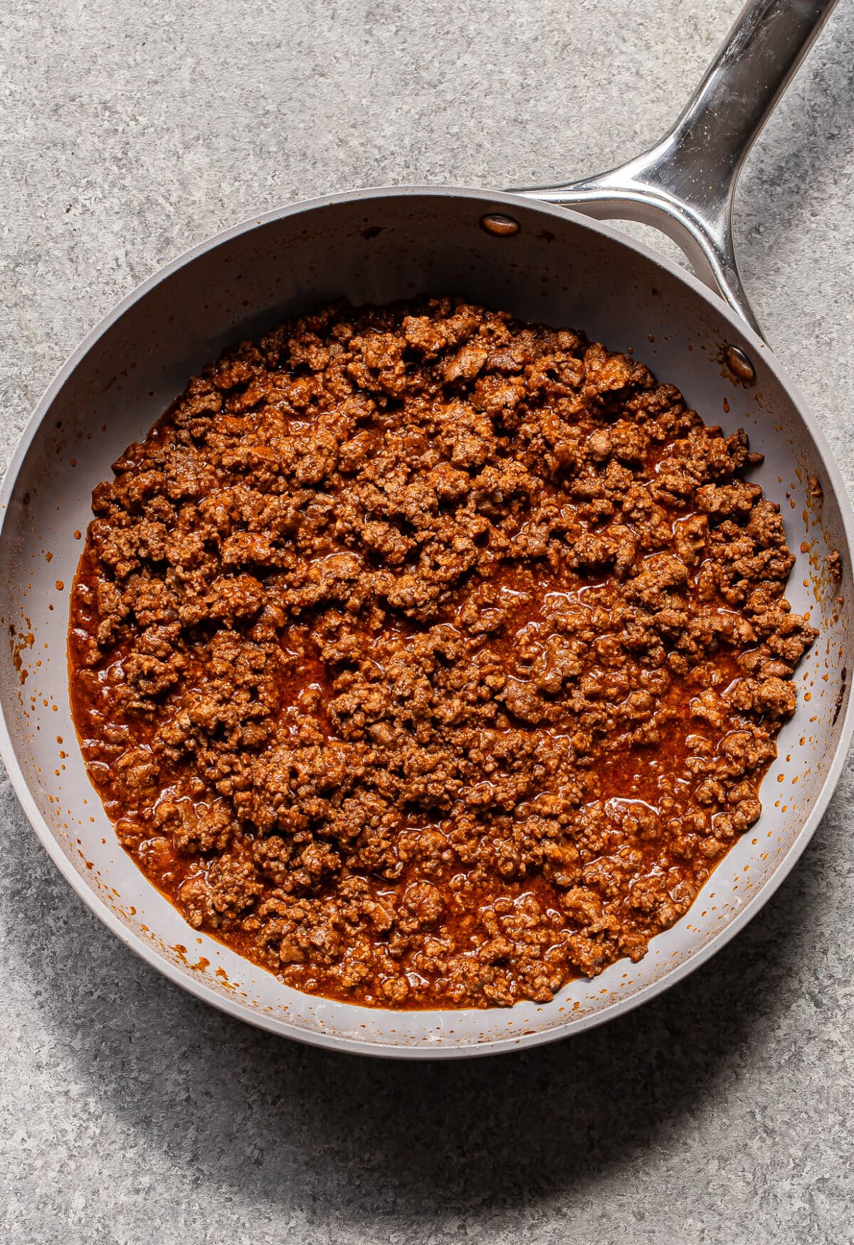 Cooked ground beef with seasoning and sauce in a silver frying pan on a gray textured surface.
