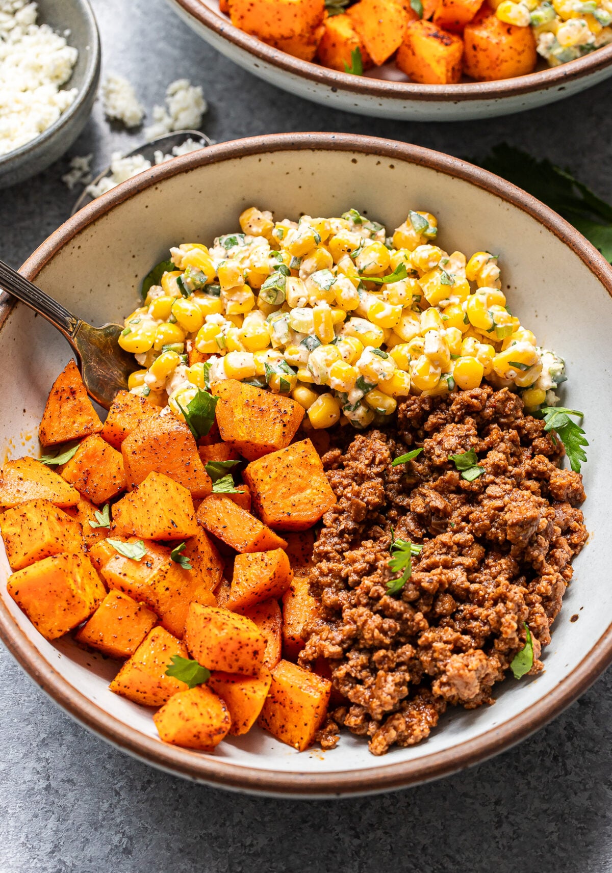 A bowl with seasoned roasted sweet potatoes, ground beef, and a corn salad with herbs and cheese, garnished with fresh parsley. A fork rests in the bowl.