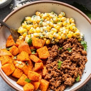 A bowl with seasoned roasted sweet potatoes, ground beef, and a corn salad with herbs and cheese, garnished with fresh parsley. A fork rests in the bowl.