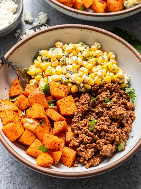 A bowl with seasoned roasted sweet potatoes, ground beef, and a corn salad with herbs and cheese, garnished with fresh parsley. A fork rests in the bowl.
