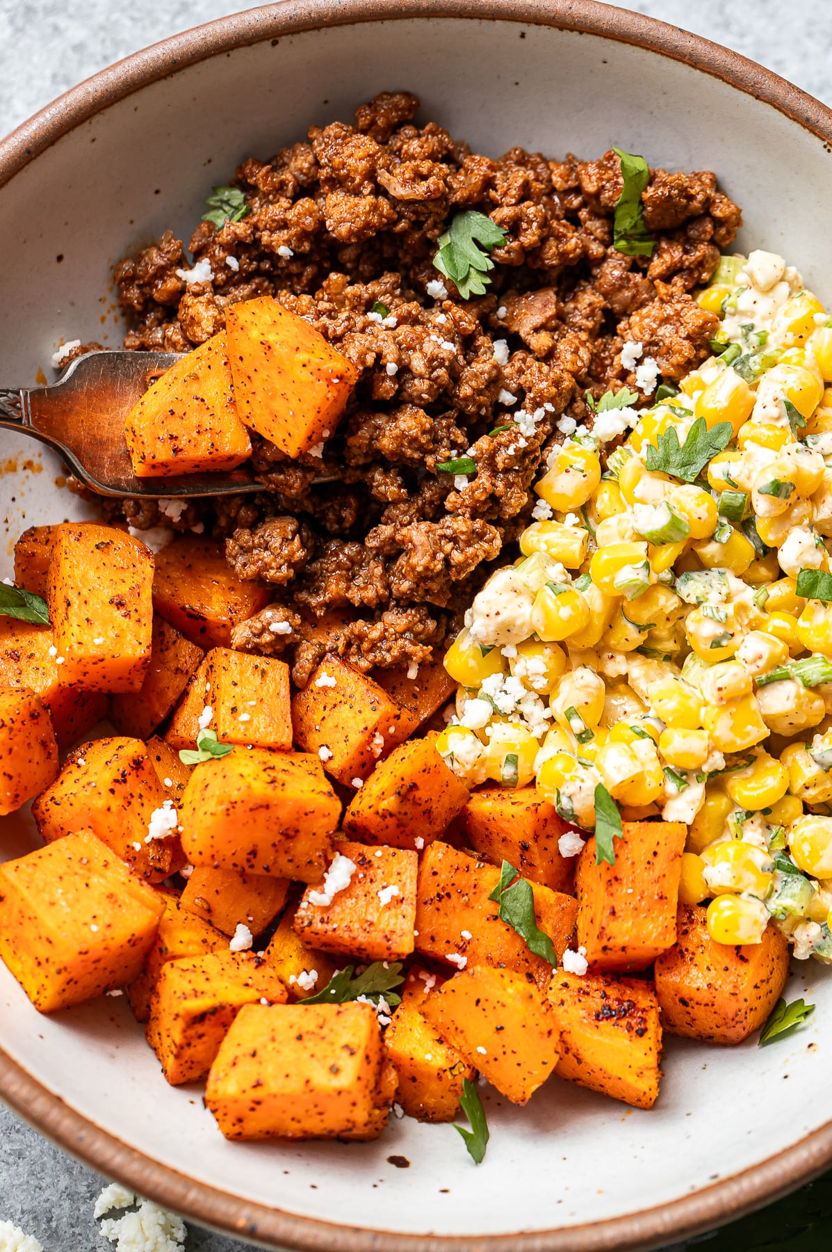 A bowl with spiced ground beef, roasted sweet potatoes, and a corn salad with cheese and herbs, garnished with parsley.
