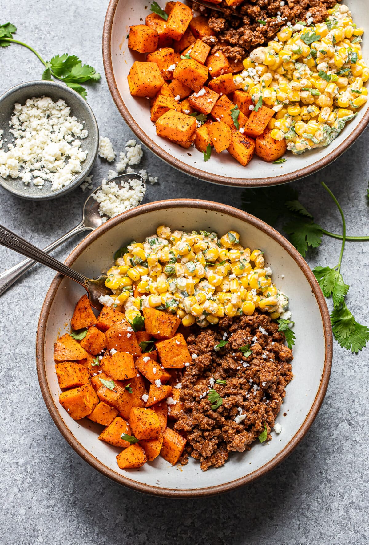 Two bowls filled with seasoned ground beef, roasted sweet potatoes, and creamy corn, topped with crumbled cheese and cilantro. A small bowl of extra cheese and scattered cilantro leaves are nearby.
