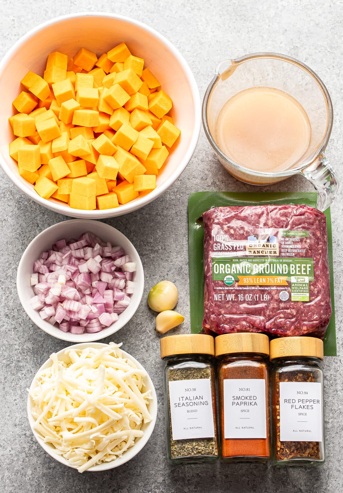 Ingredients for ground beef and butternut squash include cubed squash, diced red onion, shredded cheese, broth, organic ground beef, garlic cloves, and jars of Italian seasoning, smoked paprika, and red pepper flakes.