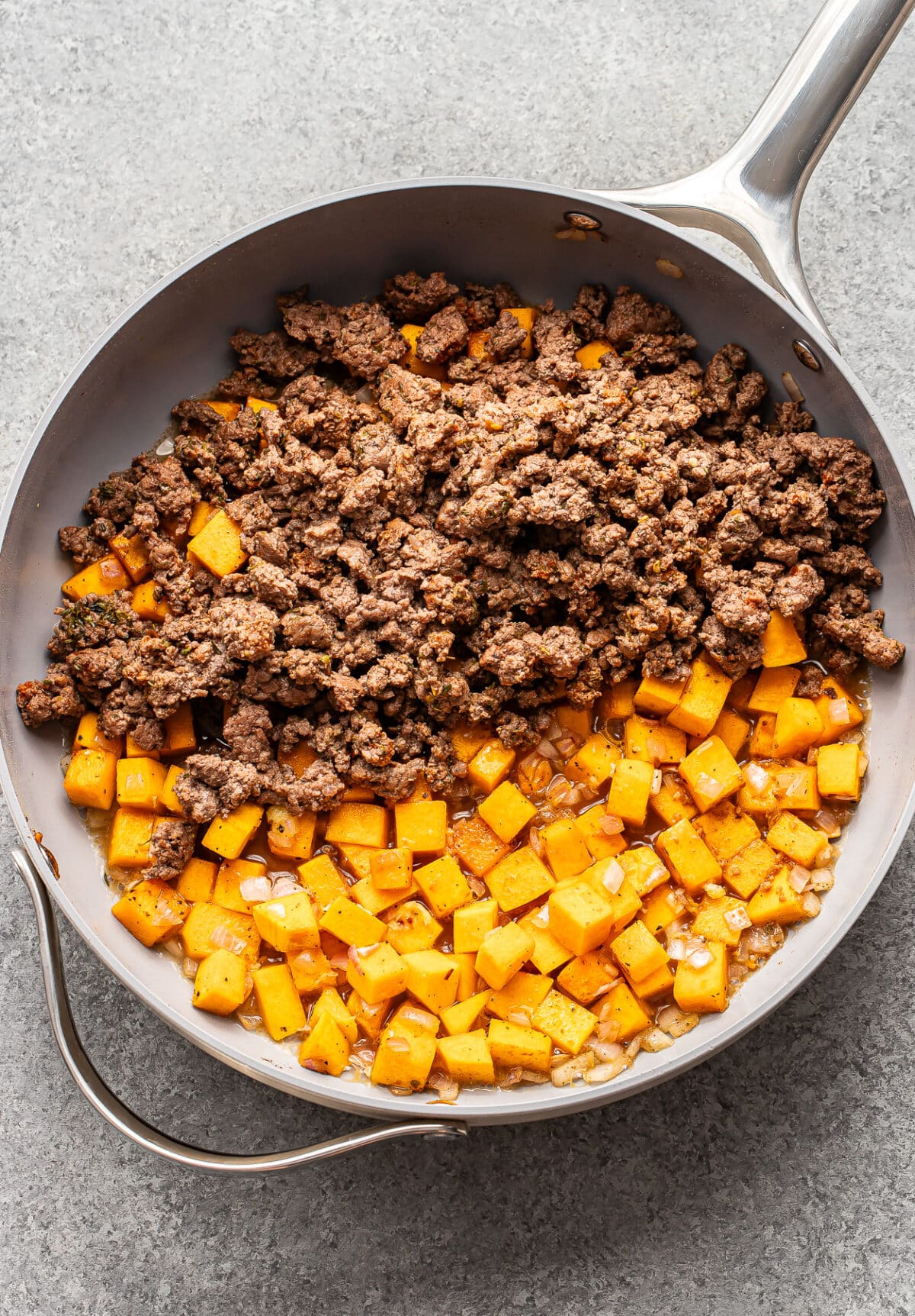 A skillet containing ground beef and butternut squash separated from a mixture of sautéed cubes and diced onions.