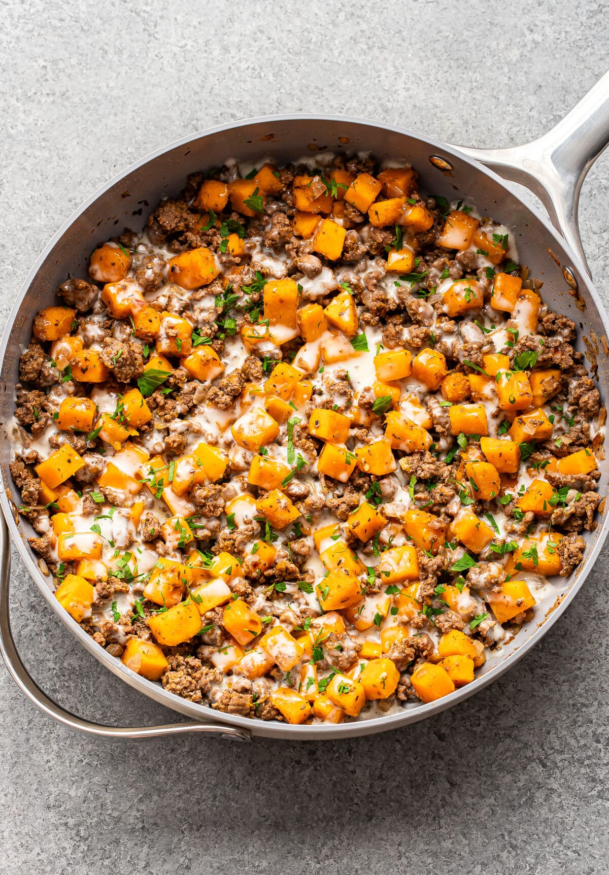 A skillet filled with ground beef and butternut squash, drizzled with a creamy sauce and sprinkled with fresh herbs.