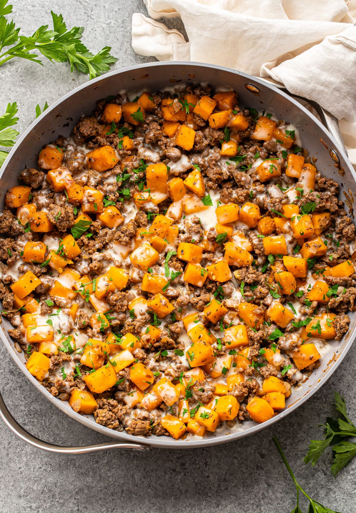 A skillet of ground beef and butternut squash topped with melted cheese and fresh herbs.