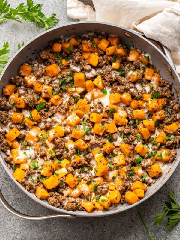 A skillet of ground beef and butternut squash topped with melted cheese and fresh herbs.