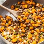 A wooden spoon stirs a skillet of sautéed butternut squash and ground beef, topped with melted cheese and a garnish of fresh herbs.
