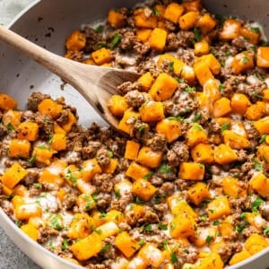 A wooden spoon stirs a skillet of sautéed butternut squash and ground beef, topped with melted cheese and a garnish of fresh herbs.
