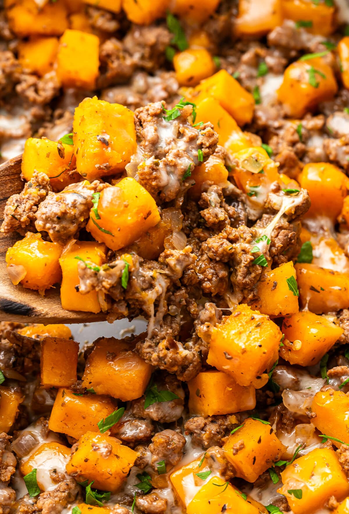 A wooden spoon scoops a mixture of ground beef and butternut squash with melted cheese, garnished with fresh herbs.