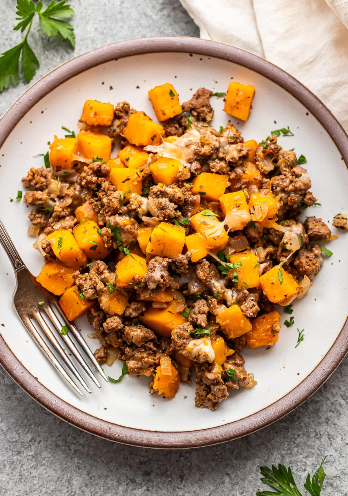 A plate of ground beef and butternut squash hash topped with melted cheese and fresh herbs, with a fork on the side.