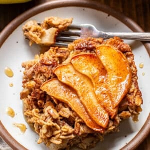 A serving of pear baked oatmeal topped with cinnamon-spiced apple slices on a small plate, with a fork lifting a piece.
