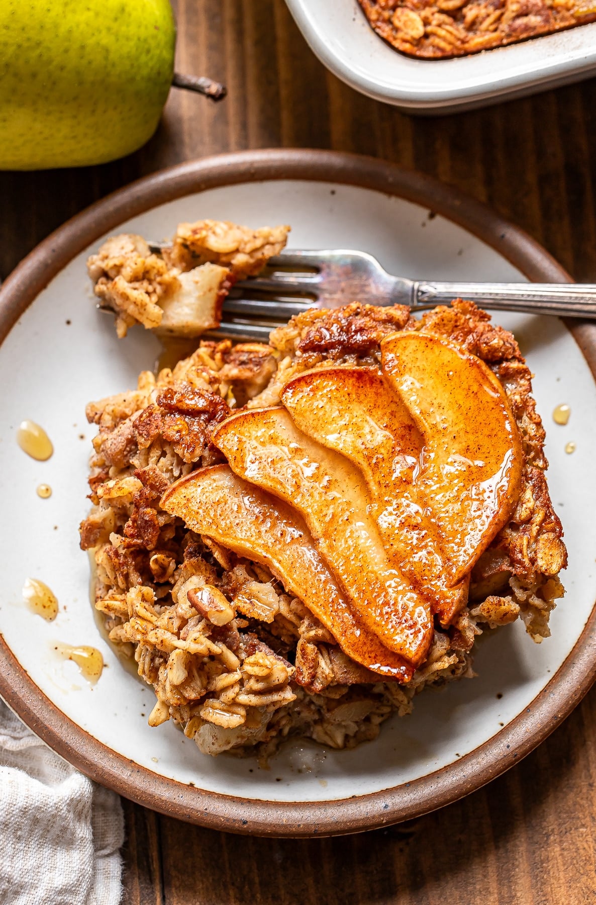 A serving of pear baked oatmeal topped with cinnamon-spiced apple slices on a small plate, with a fork lifting a piece.