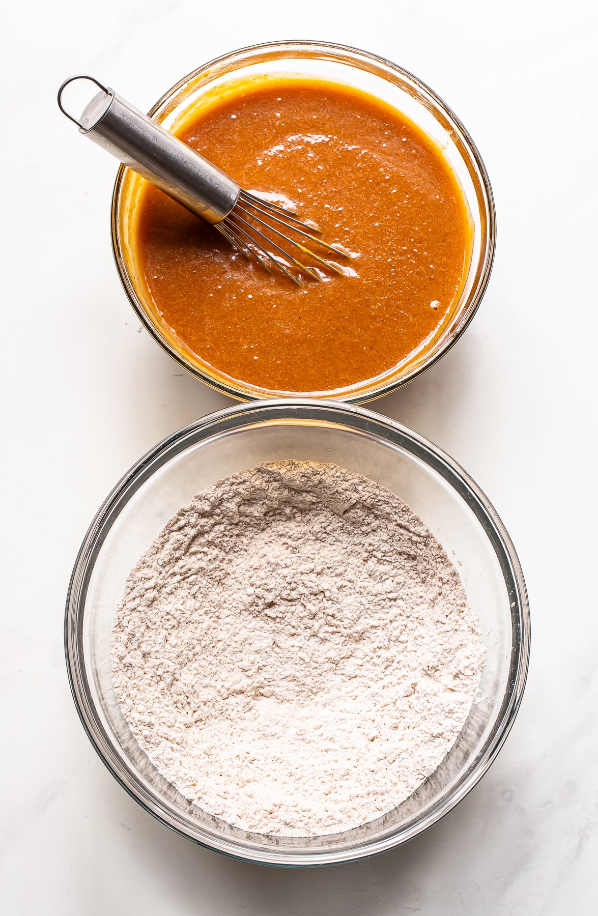 Two glass bowls, one containing a whisked orange batter for pumpkin bread and the other containing dry flour, sit on a white surface.
