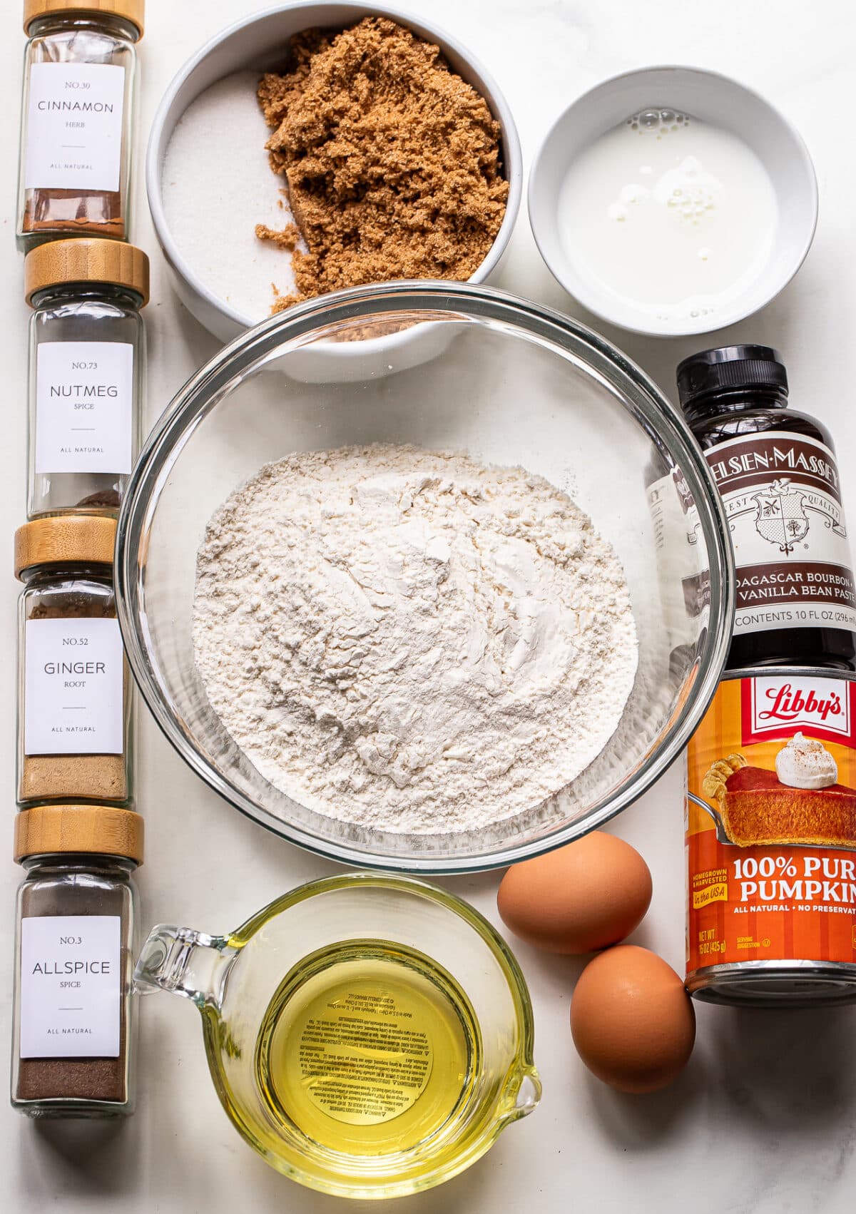 A glass bowl of flour surrounded by jars of spices, brown sugar, white sugar, milk, oil, eggs, pumpkin puree, and vanilla bean paste for baking pumpkin bread.