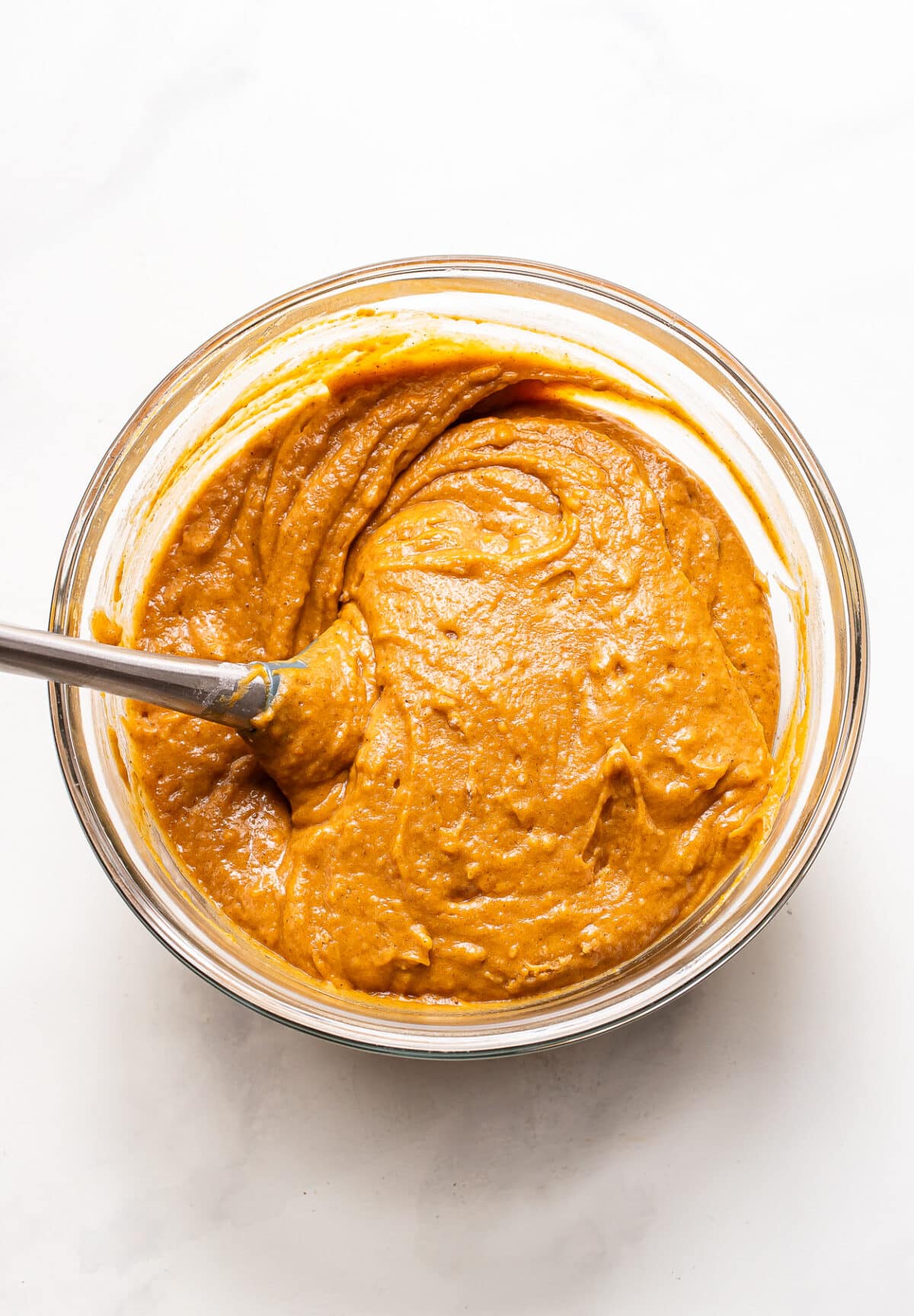 A metal spoon stirs thick, orange pumpkin bread batter in a glass mixing bowl.