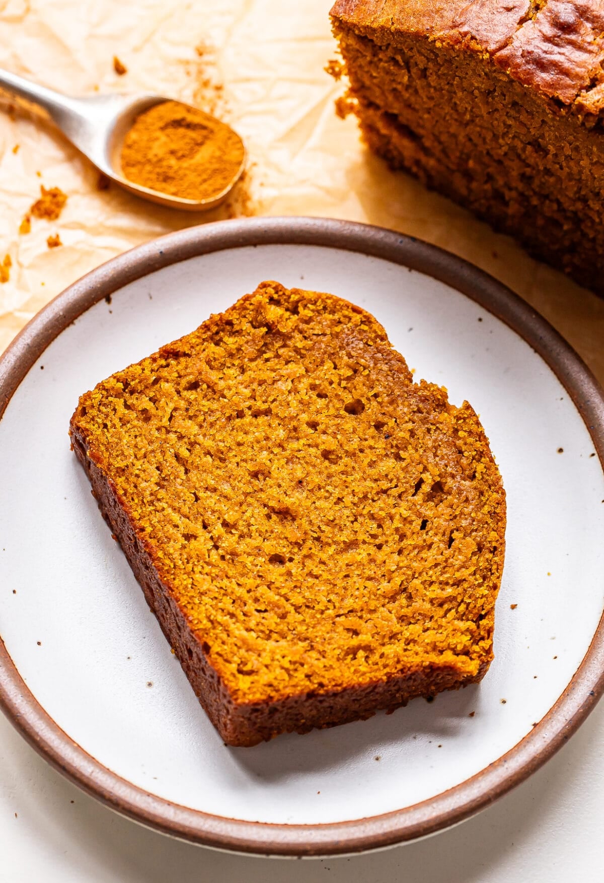 A slice of pumpkin bread on a small plate, with a loaf of the bread and a spoonful of spice in the background.