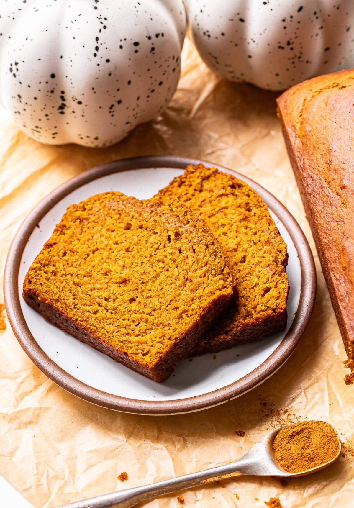 Two slices of pumpkin bread on a plate next to a loaf, with a spoon of ground spice nearby.