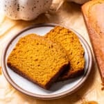 Two slices of pumpkin bread on a plate next to a loaf, with a spoon of ground spice nearby.