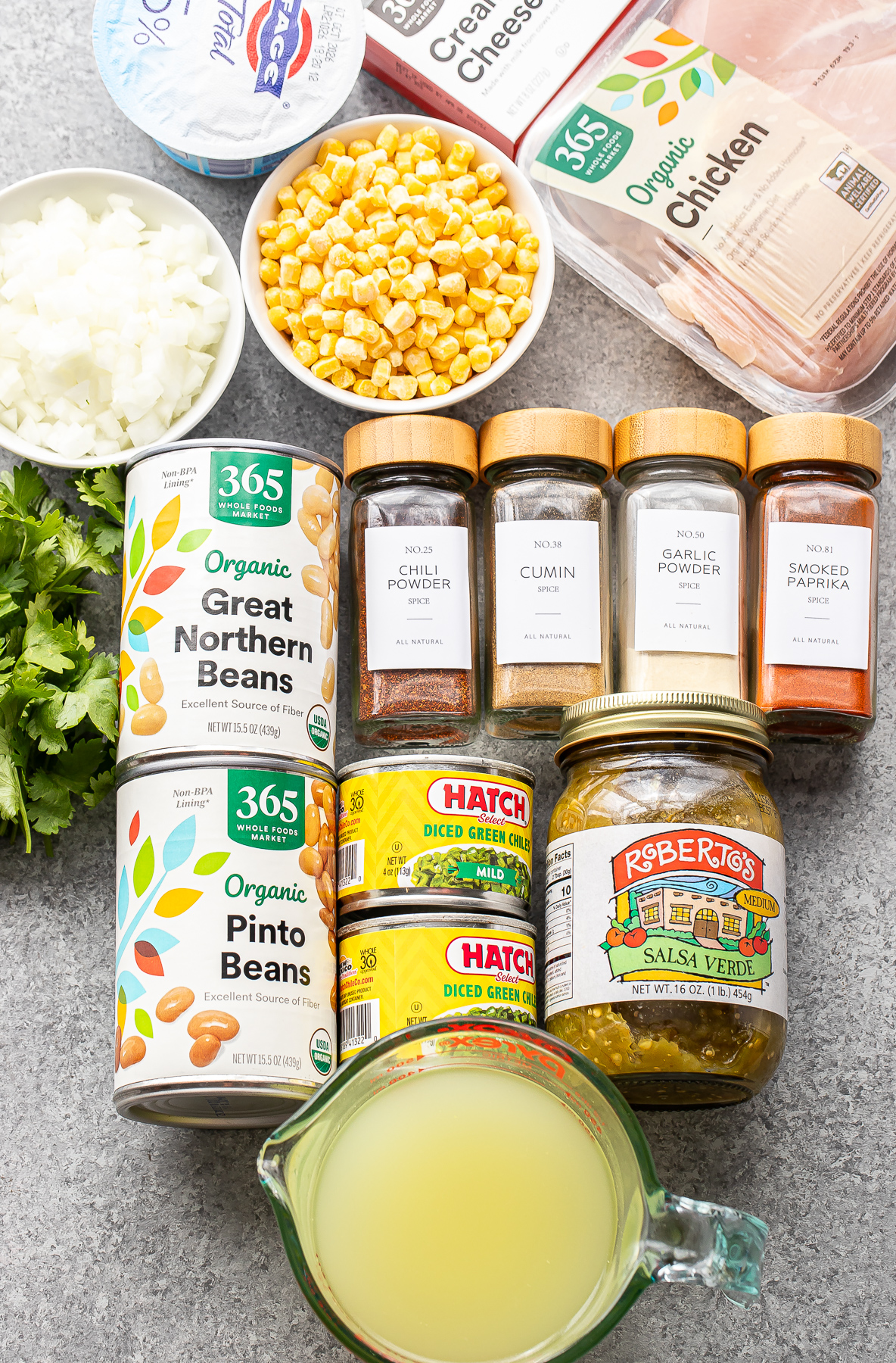 Ingredients for a recipe arranged on a gray surface, including chicken breast, corn, onions, cilantro, canned beans, diced green chiles, salsa verde, a measuring cup of broth, and various dried spices.