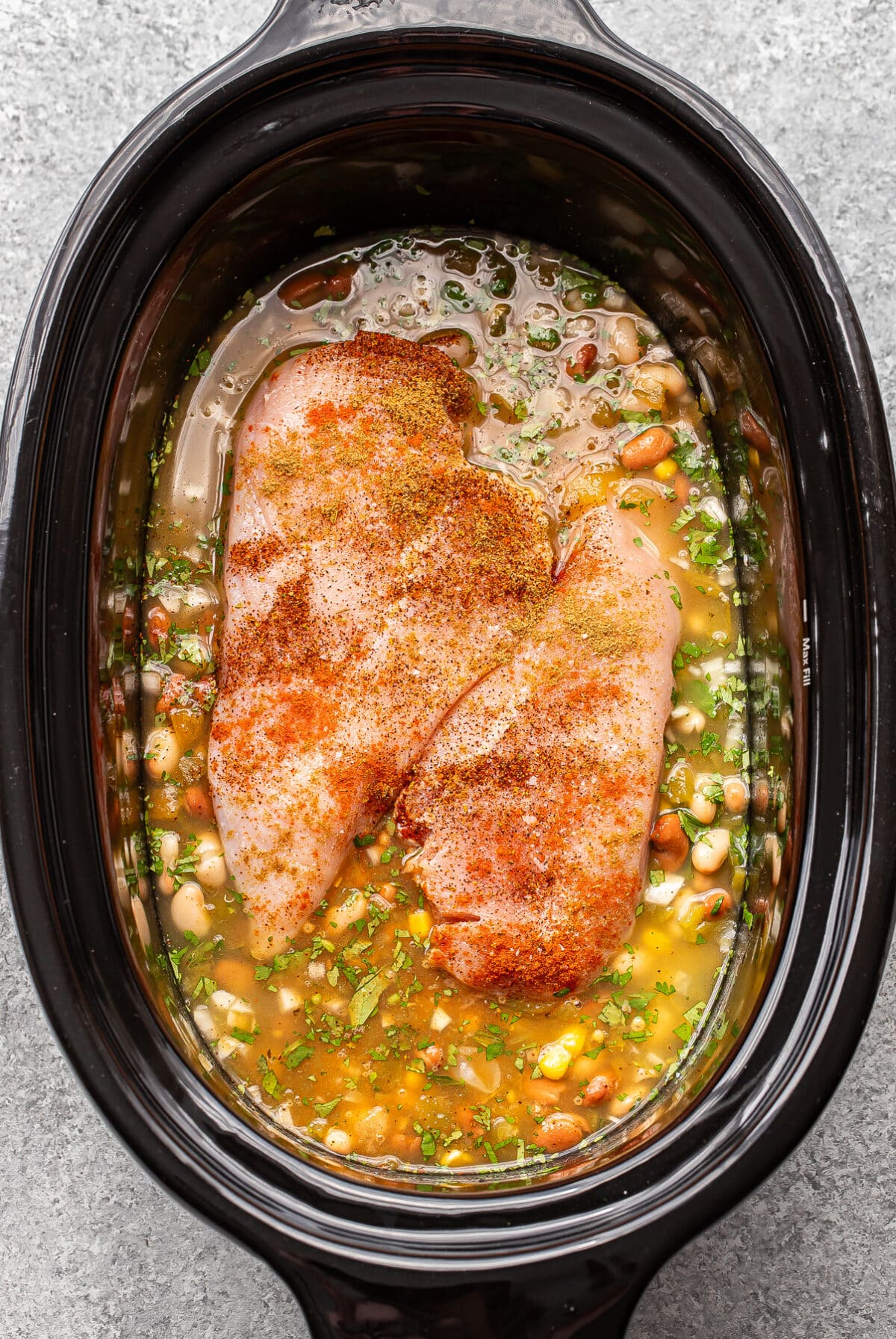 Two raw seasoned chicken breasts resting in a slow cooker filled with beans, broth, and herbs.