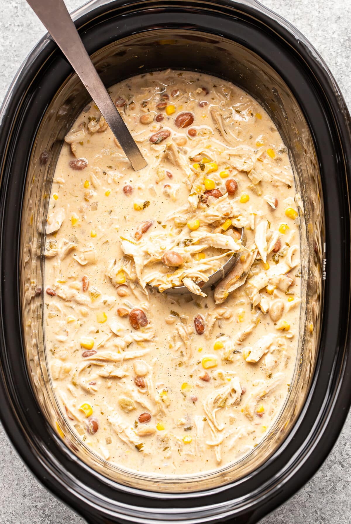 A slow cooker filled with creamy chicken chili containing corn, beans, and shredded chicken, with a metal ladle partially submerged in the center.
