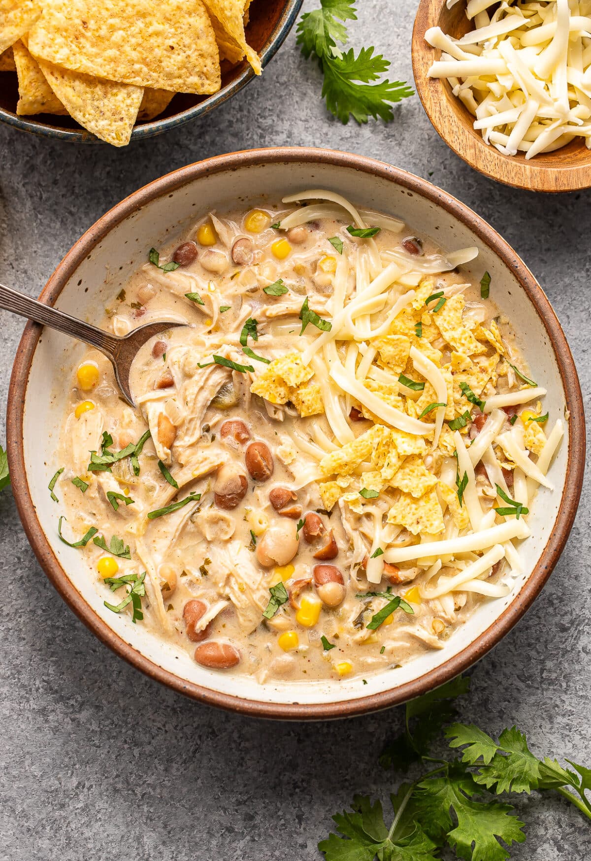 A bowl of creamy white chicken chili topped with shredded cheese and tortilla chips, with a spoon inside and side bowls of chips and cheese nearby.