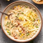 A bowl of creamy white chicken chili topped with shredded cheese and tortilla chips, with a spoon inside and side bowls of chips and cheese nearby.