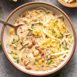 A bowl of creamy white chicken chili topped with shredded cheese and tortilla chips, with a spoon inside and side bowls of chips and cheese nearby.