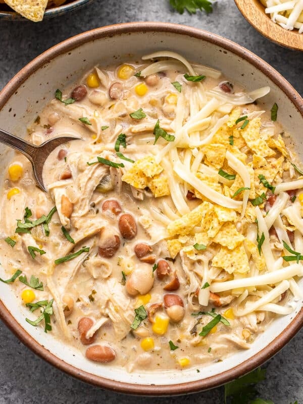 A bowl of creamy white chicken chili topped with shredded cheese, tortilla chips, and fresh herbs, with a spoon inside.